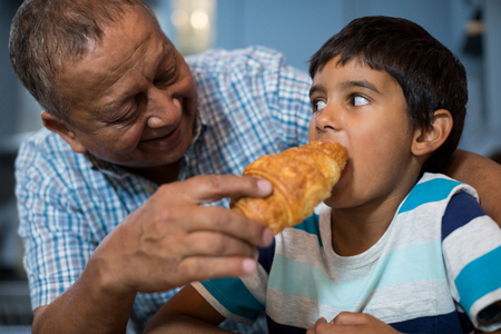 Grandfather feeding croissant to grandson while having breakfastの写真素材