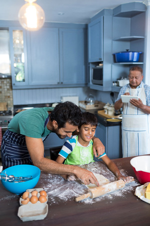 Father assisting son for rolling dough with man using tablet in backgroundの写真素材