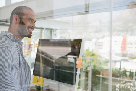 Smiling designer looking away while standing by computer seen through glassの写真素材