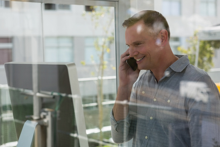 Smiling fashion designer talking on mobile phone by computer seen through glassの写真素材