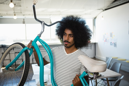 Young man with curly hair holding bicycle at creative officeの写真素材