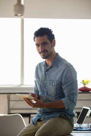 Portrait of smiling man using tablet while sitting on desk in officeの写真素材