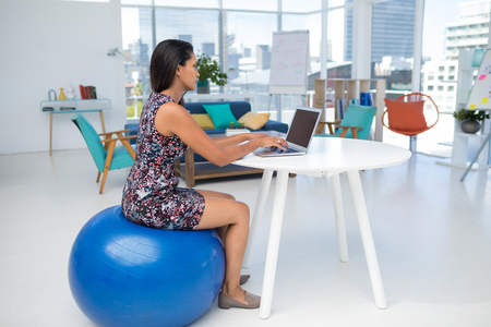 Female Executive Using Laptop While Sitting On Exercise Ball At Executive Using Laptop While Sitting
