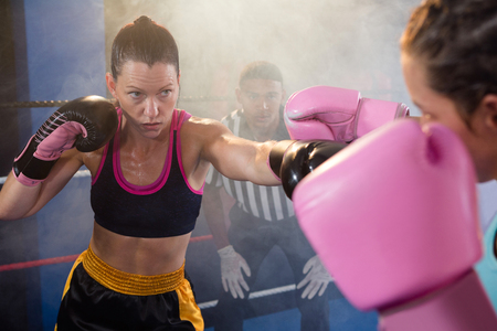 Female athletes fighting while referee looking in boxing ring at fitness studioの写真素材