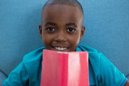 Close-up portrait of smiling boy with red novel sitting on armchair at homeの写真素材