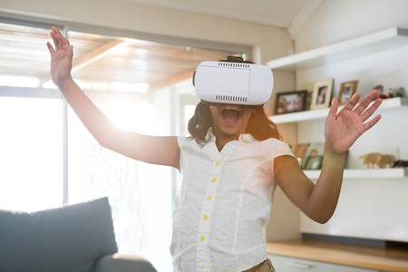 Cheerful girl wearing virtual reality simulator while standing in living room at homeの写真素材
