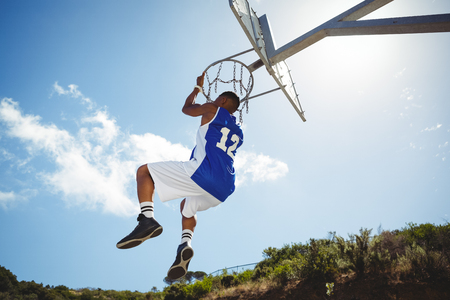 Rear view of man hanging on basketball hoop against blue skyの写真素材