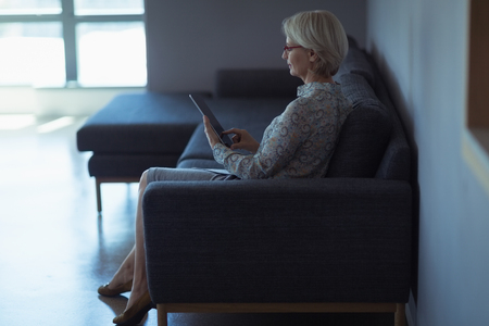 Side view of businesswoman using digital tablet while sitting on sofa in officeの写真素材