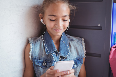 Elementary schoolgirl listening music through headphones while using mobile phone by wall at schoolの写真素材