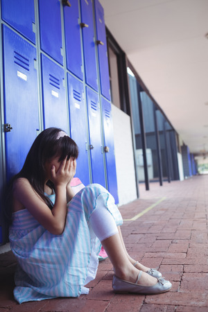 Side view of schoolgirl covering face with hands while sitting by lockers at schoolの写真素材