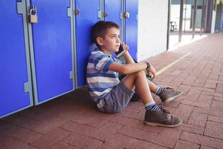 High angle view of boy talking on mobile phone while sitting by lockers in corridor at schoolの写真素材