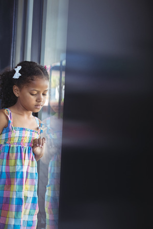 Thoughtful girl looking down while standing by window in schoolの写真素材