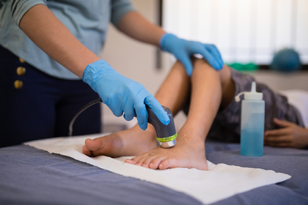 Female therapist wearing gloves scanning feet of boy lying on bed at hospital wardの写真素材