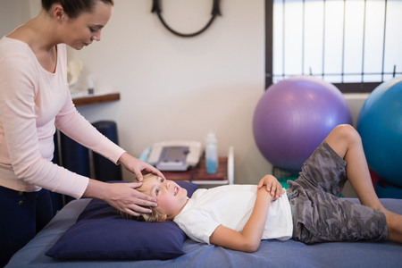 Young female therapist giving head massage to boy lying on bed at hospital wardの写真素材
