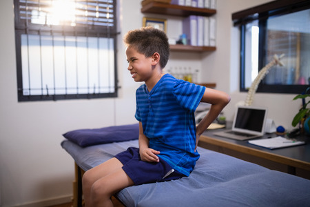 Boy frowning with backache while sitting on bed at hospital ward
