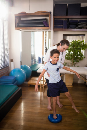 Female therapist holding boy standing on blue stress ball at hospital wardの写真素材