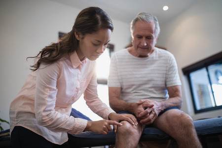 Low angle view of female therapist examining knee while male patient sitting on bed at hospital wardの写真素材