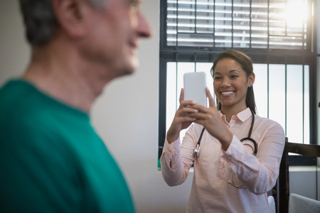 Smiling female therapist photographing senior male patient against window at hospital wardの写真素材