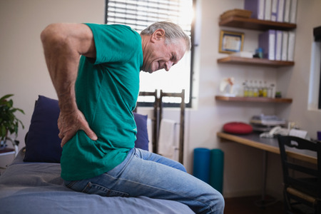 Side view of male patient grimacing from back ache while sitting on bed at hospital ward
