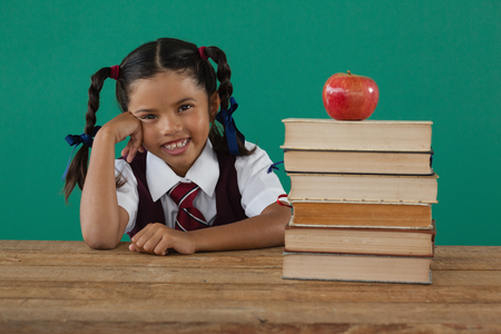 Portrait of schoolgirl sitting beside books stack with apple on top against chalkboardの写真素材