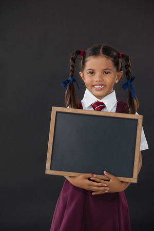 Portrait of schoolgirl holding blank slate against blackboard in classroomの写真素材