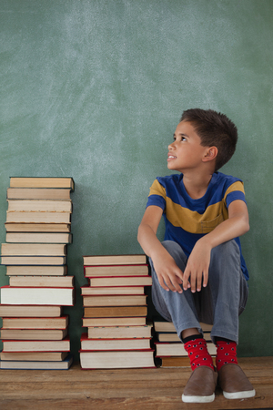 Thoughtful schoolboy sitting on books stack against chalkboardの写真素材