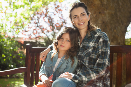 Portrait of smiling beautiful woman sitting with daughter on wooden bench at backyardの写真素材