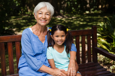 Portrait of senior woman with granddaughter sitting on wooden bench at backyardの写真素材