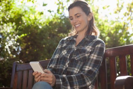 Low angle view of smiling beautiful woman text messaging while sitting on wooden bench at backyardの写真素材