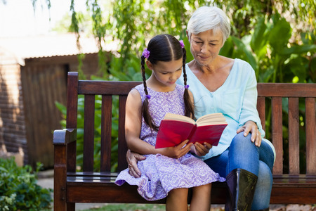 Senior woman sitting by girl reading book on wooden bench at backyard