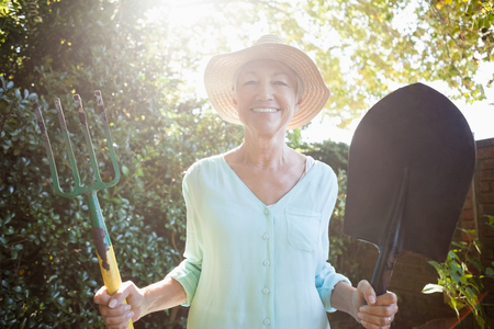 Back lit portrait of smiling senior woman holding garden fork and shovel at backyardの写真素材