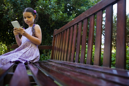 Surface level of girl sitting on wooden bench while using mobile phone at backyardの写真素材