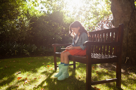 Smiling girl using smart phone while sitting on wooden bench in backyard during sunny dayの写真素材