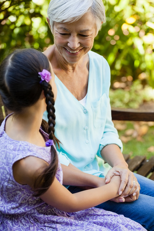 Smiling grandmother holding hands of granddaughter while sitting on bench at backyardの写真素材