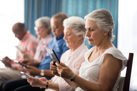Row of senior people sitting on chairs using digital tablets at retirement homeの写真素材