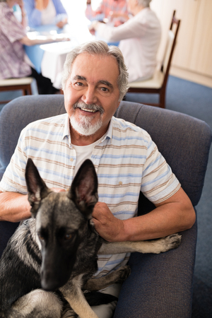 Portrait of smiling senior man sitting with puppy on armchair at retirement homeの写真素材