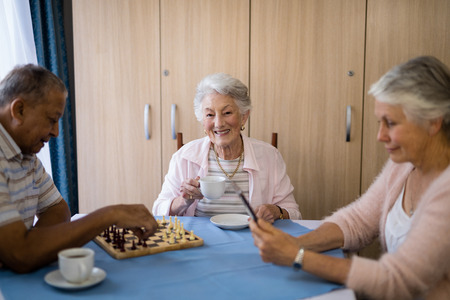 Smiling woman having coffee while sitting with friends playing chess and using technology at table in nursing homeの写真素材
