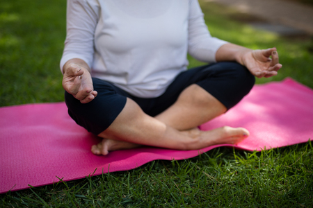 Mid section of senior woman meditating while sitting on exercise mat at parkの写真素材