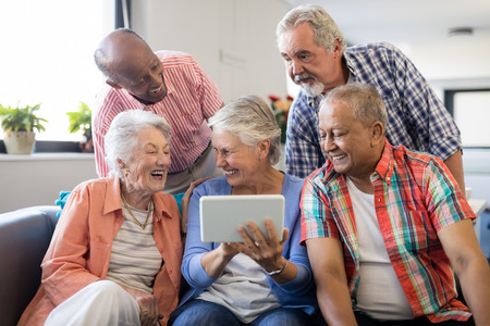 Senior woman showing digital tablet to cheerful friends while sitting on sofa at nursing homeの写真素材