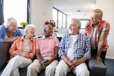 Smiling senior friends listening to man while sitting on sofa in nursing homeの写真素材