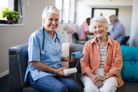 Portrait of senior woman with female doctor holding blood pressure gauge while sitting on sofa at homeの写真素材