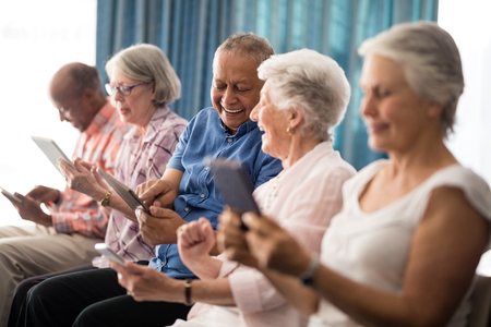 Smiling senior people sitting on chairs while using digital tablets at retirement homeの写真素材