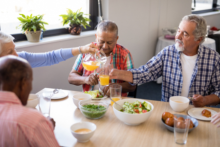 Senior woman serving drink to friends while having food at table in nursing homeの写真素材
