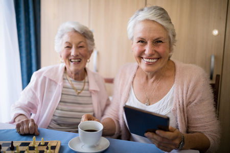 Portrait of smiling senior friends having coffee while playing chess at table in nursing homeの写真素材