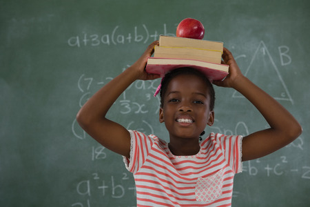 Portrait of schoolgirl holding books stack with apple on head against chalkboardの写真素材