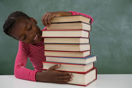 Happy schoolgirl sitting with books stack against chalkboardの写真素材