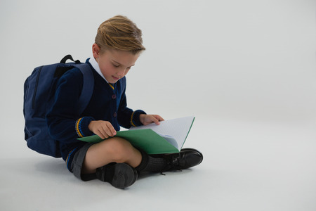 Adorable schoolboy reading book while sitting on white backgroundの写真素材
