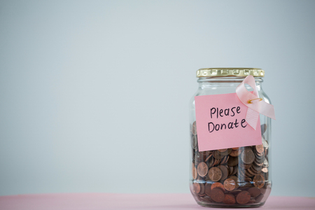 Close-up of card and pink Breast Cancer Awareness ribbon on jar with coins on table against white backgroundの写真素材