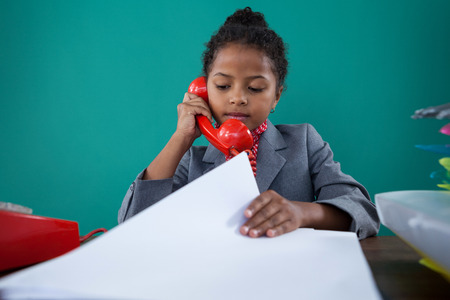 Kid Businesswoman checking file while talking on land line at desk against blue backgroundの写真素材