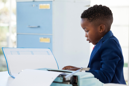 Side view of kid businessman reading document while working at desk in officeの写真素材